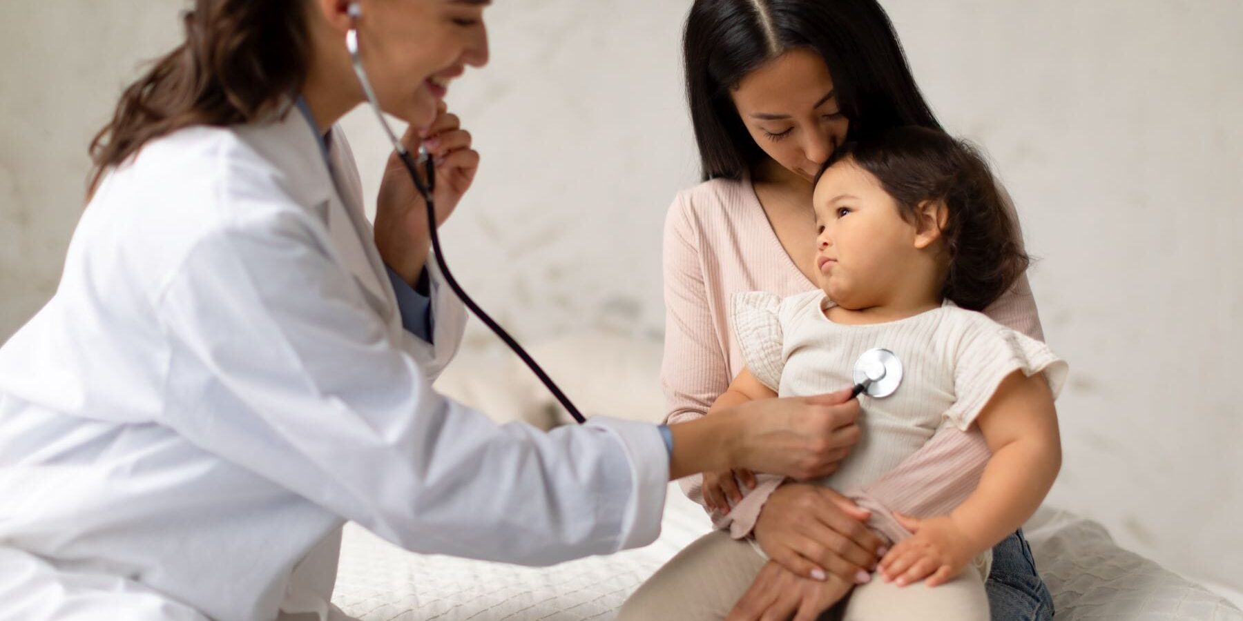 Pediatrician Lady Listening To Korean Infant Child's Heartbeat Pressing Stethoscope To Little Girl's Chest During Medical Checkup Indoors. Mom And Toddler Daughter Visiting Clinic. Selective Focus